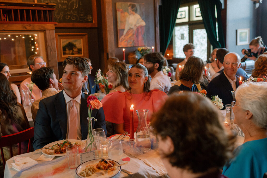Guests seated at tables decorated with orange tapered candles and vibrant florals are photographed at a wedding reception held at snapdragon vashon