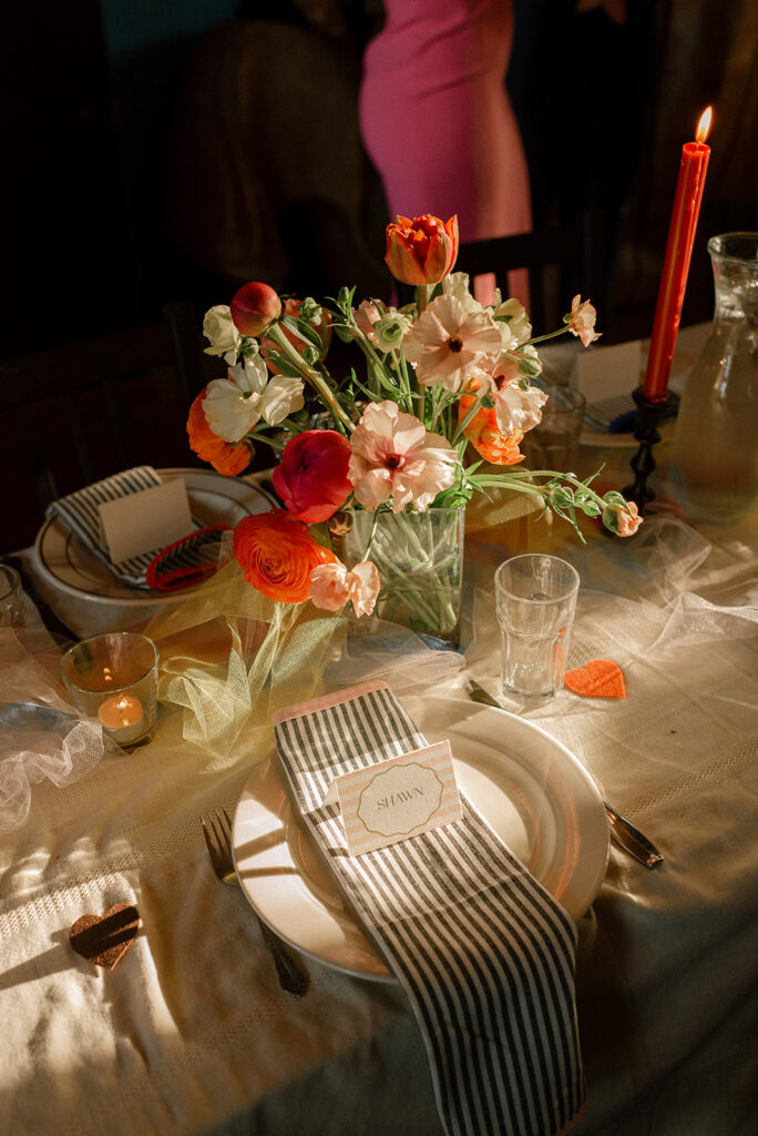 A tablescape featuring vibrant florals, deep orange tapered candles, paper confetti hearts, tulle, and china are photographed at a wedding reception held at snapdragon vashon