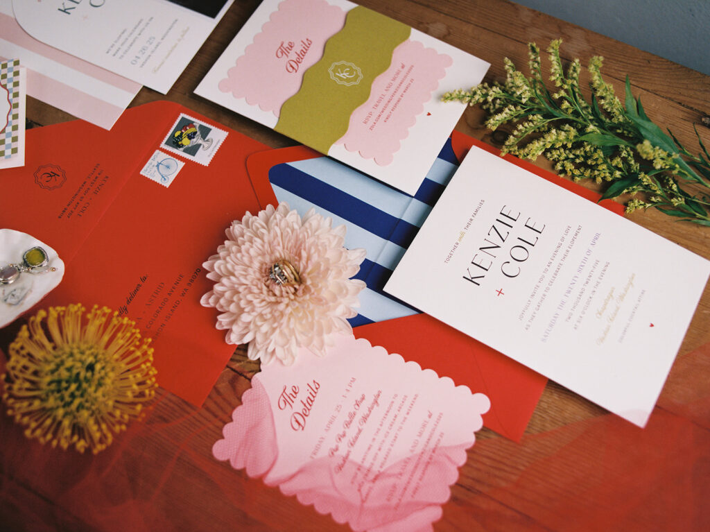 A flat lay of wedding invitations, announcements, and wedding rings atop wedding florals in shades of orange, pink, green, and blue taken by WA state wedding photographers