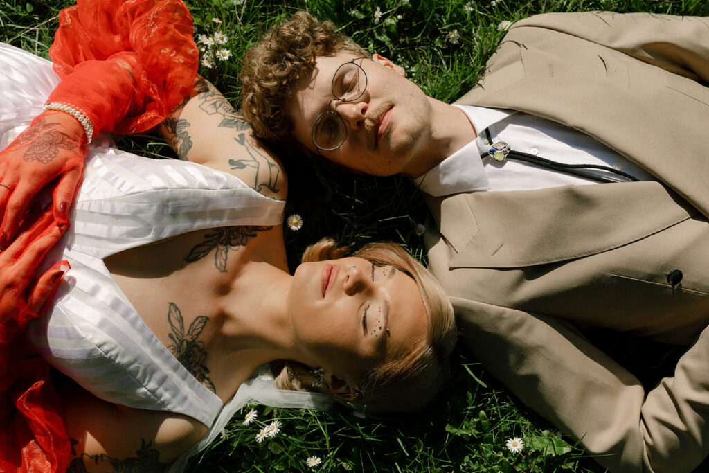 A bride wearing orange gloves lies in the grass next to her groom in a tan suit & bolo tie in a photo taken from above by washington state wedding photographers