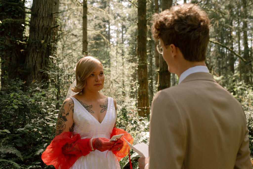 A bride wearing a white dress and orange gloves listens as her groom recites his handwritten vows during their Vashon Island wedding 