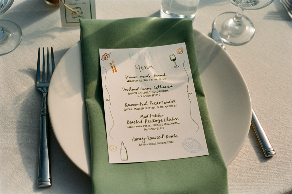 A place setting featuring a menu atop a sage green linen napkin on a white plate is displayed at a wedding reception at Captain Whidbey Inn 
