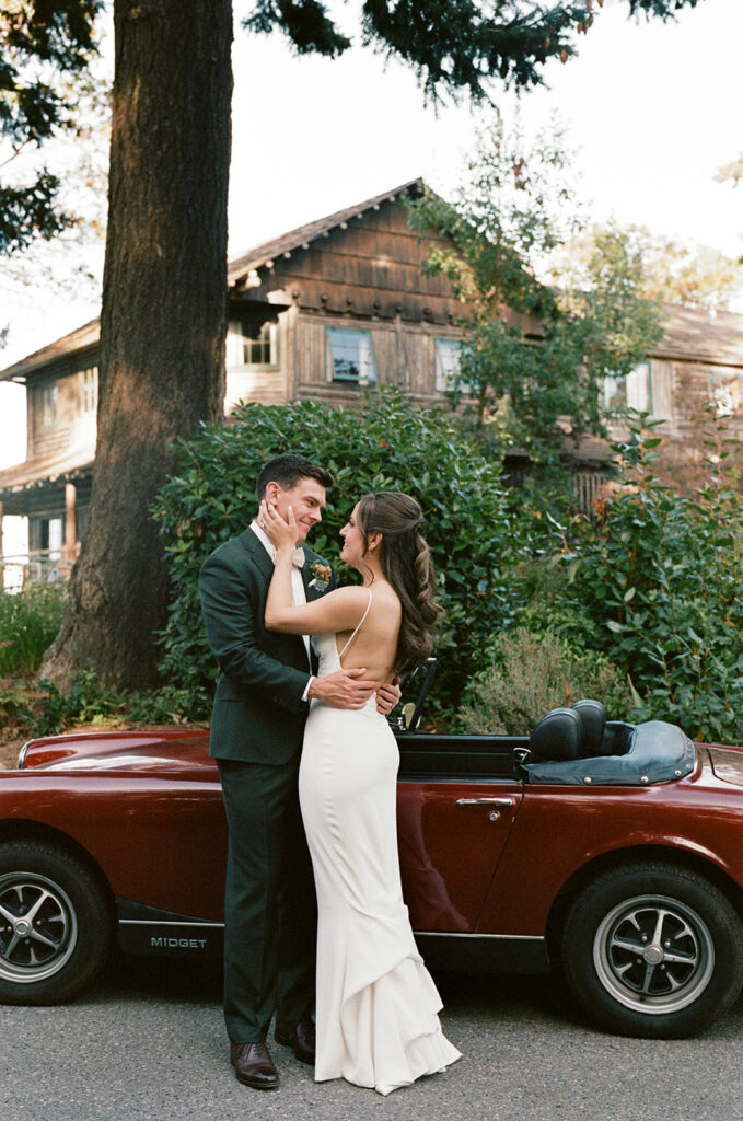 A bride and groom embrace in their wedding attire next to a maroon vintage convertible at their Captain Whidbey Inn wedding 