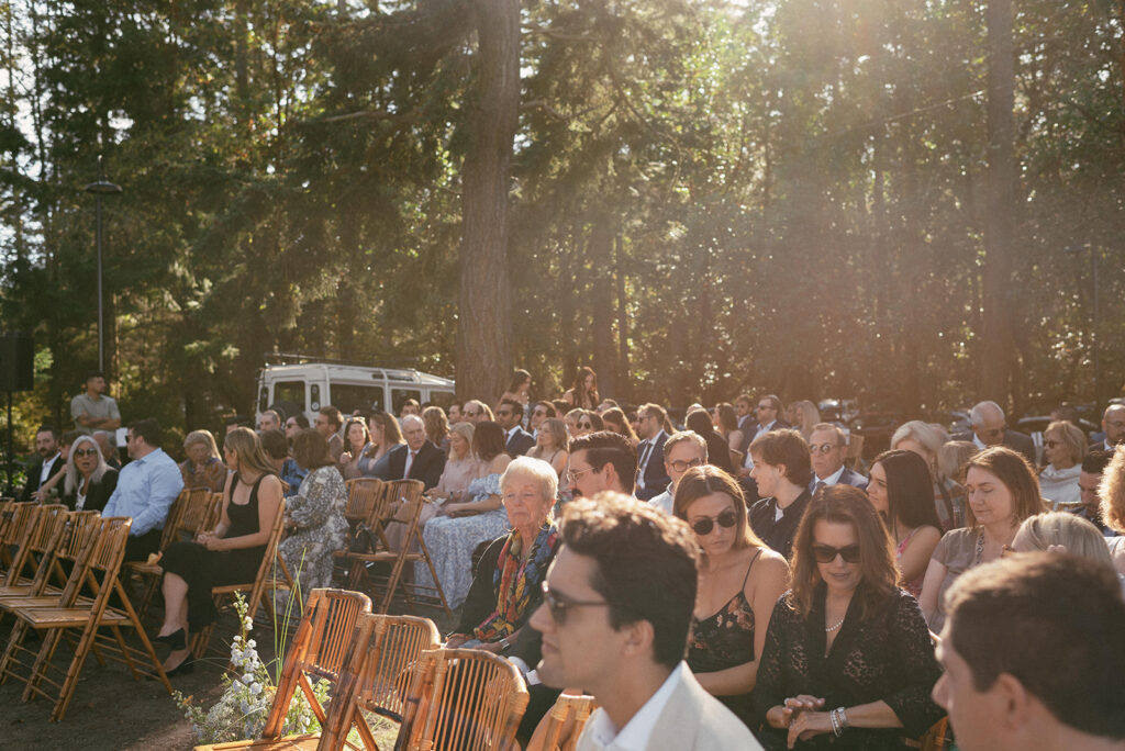 Guests are seated in wooden chairs at an outdoor event space awaiting a Captain Whidbey Inn wedding ceremony