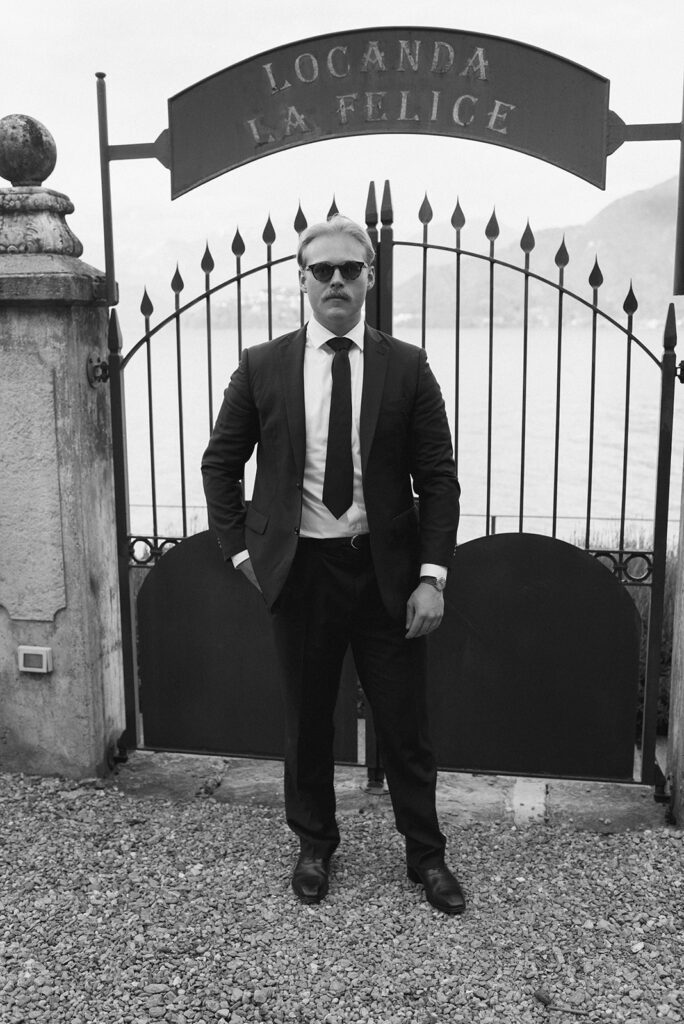 A groom wearing a black suit stands before an iron gate in a b+w image taken at his destination wedding on Lake Como 