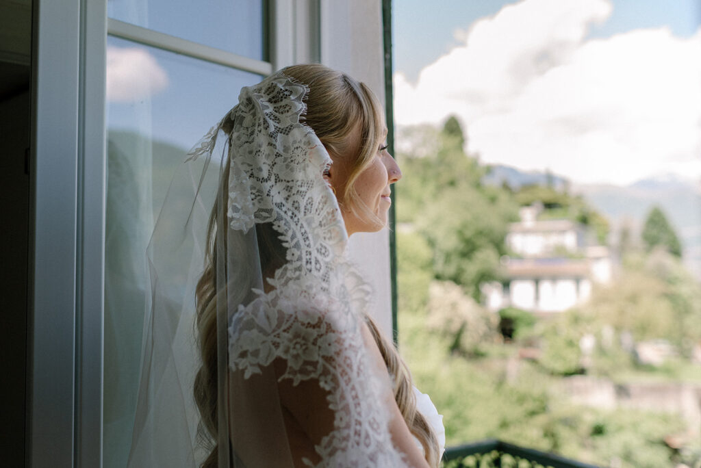 A bride wearing a lace veil looks out over the water from her room at Villa la Felice before her destination wedding on Lake Como 