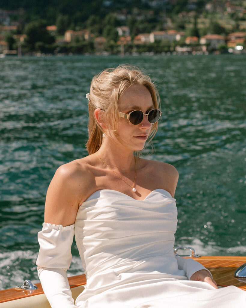 A bride relaxes on a boat wearing sunglasses & her wedding dress following their destination wedding on Lake Como 
