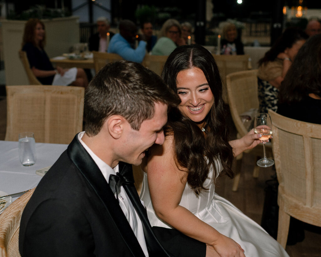 Newlyweds laugh seated next to each other in a documentary style photo taken at their reception at their greenhouse wedding venue mn
