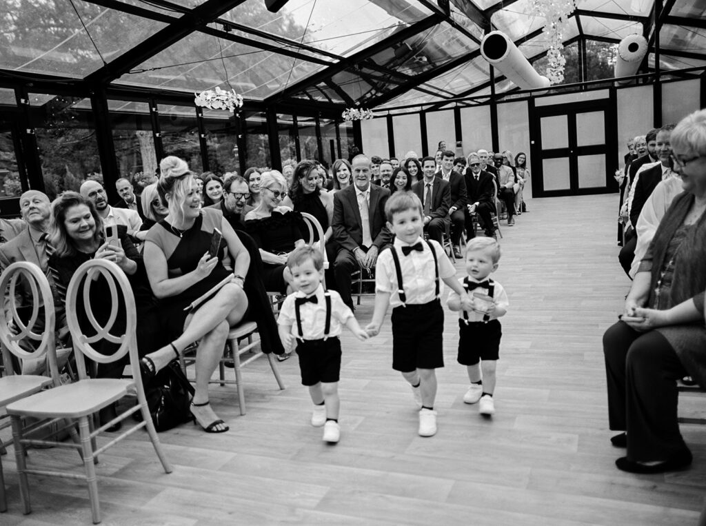 Three small boys walk hand-in-hand down the aisle of the ceremony atrium at Woodland GLasshaus,  chaska mn wedding venues 