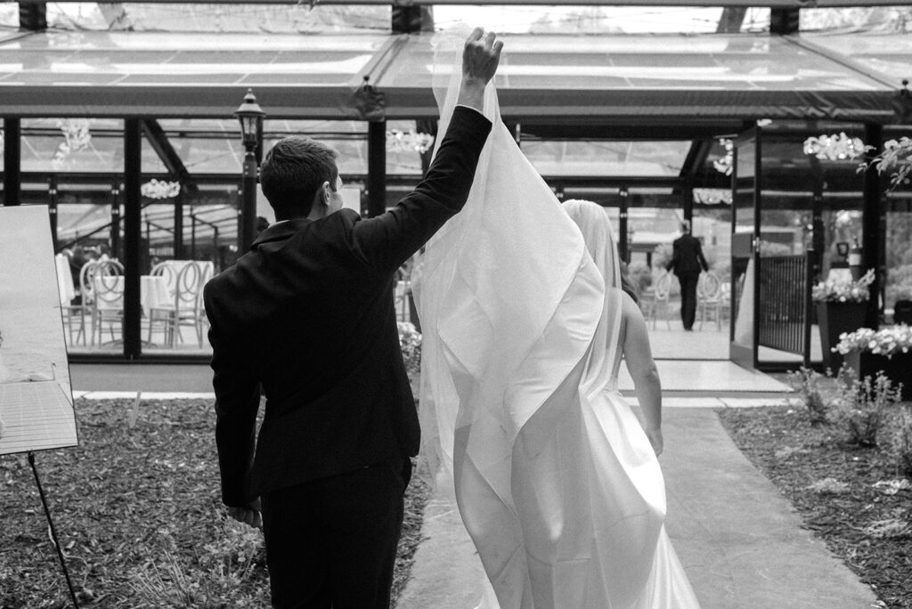 A groom assists his bride with her veil as they walk into the dinner atrium at Woodland Glasshaus, one of top outdoor wedding venues minnesota