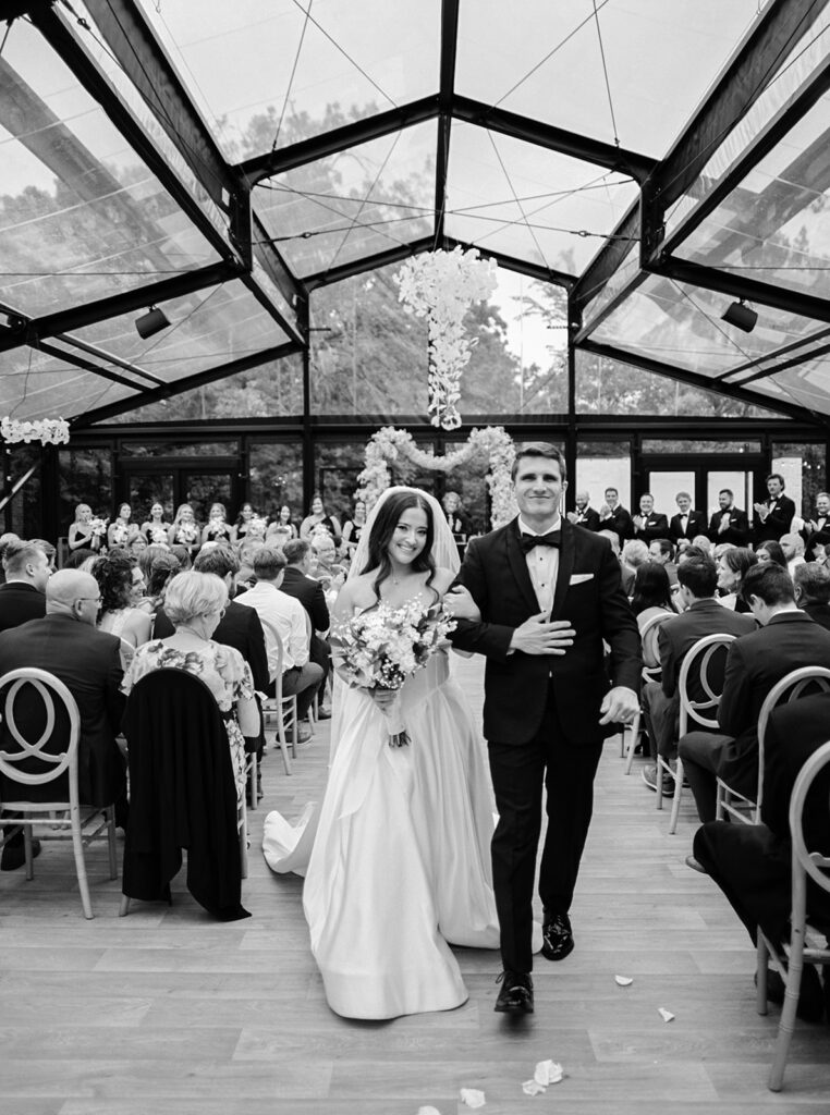 A black and white image depicts newlyweds walking down the aisle in the ceremony atrium at a venue space in bavaria downs chaska