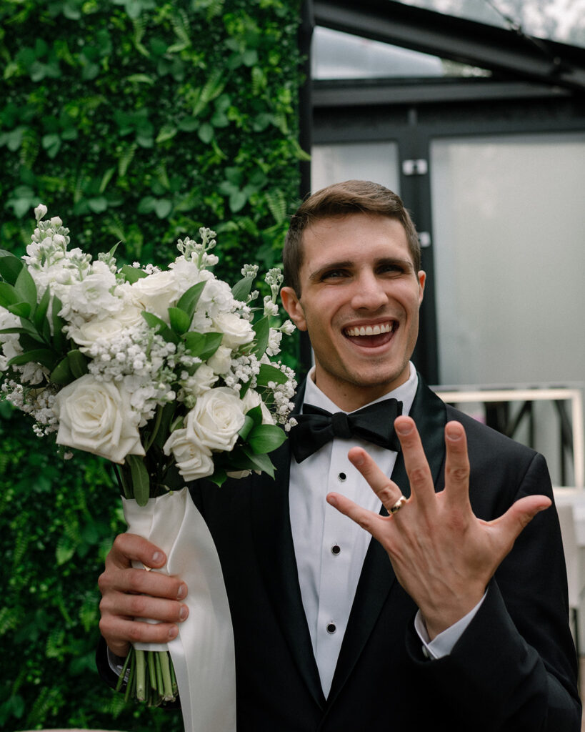 A groom in a black tux shows off his golden wedding band following his ceremony at a greenhouse wedding venue mn