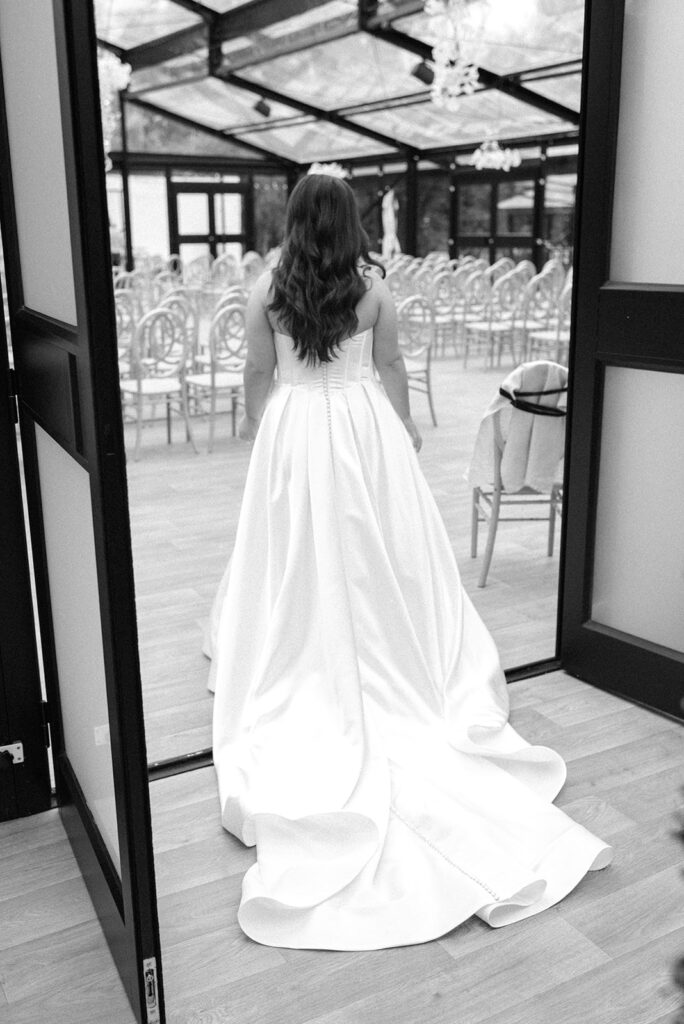 A b+w image depicts a bride in a strapless wedding gown looking into the ceremony atrium at her Bavarian Downs wedding at Woodland Glasshaus
