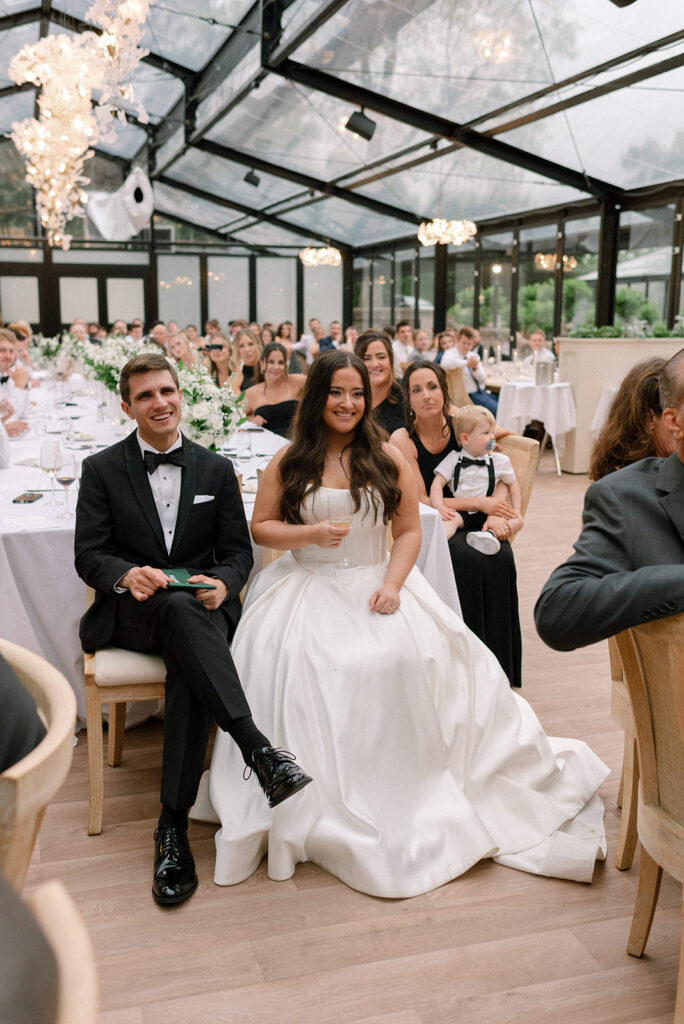Newlyweds smile as they listen to speeches at their reception in the dining atrium at Woodland Glasshaus, a chaska mn wedding venues