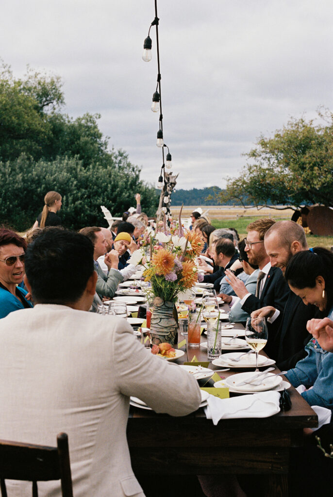 Wedding guests laugh & chat at a long dinner table covered in florals & drinks at a Tokeland Hotel wedding in Tokeland Washington 