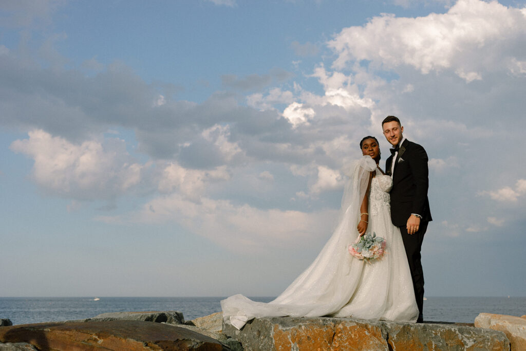 A bride & groom pose in their wedding attire on the coast of their New England wedding at Barker House by Wedgewood Weddings 