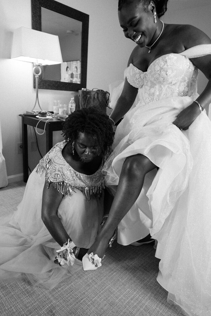 The mother of the bride assists the bride with her shoes in a black and white image taken before her New England wedding 