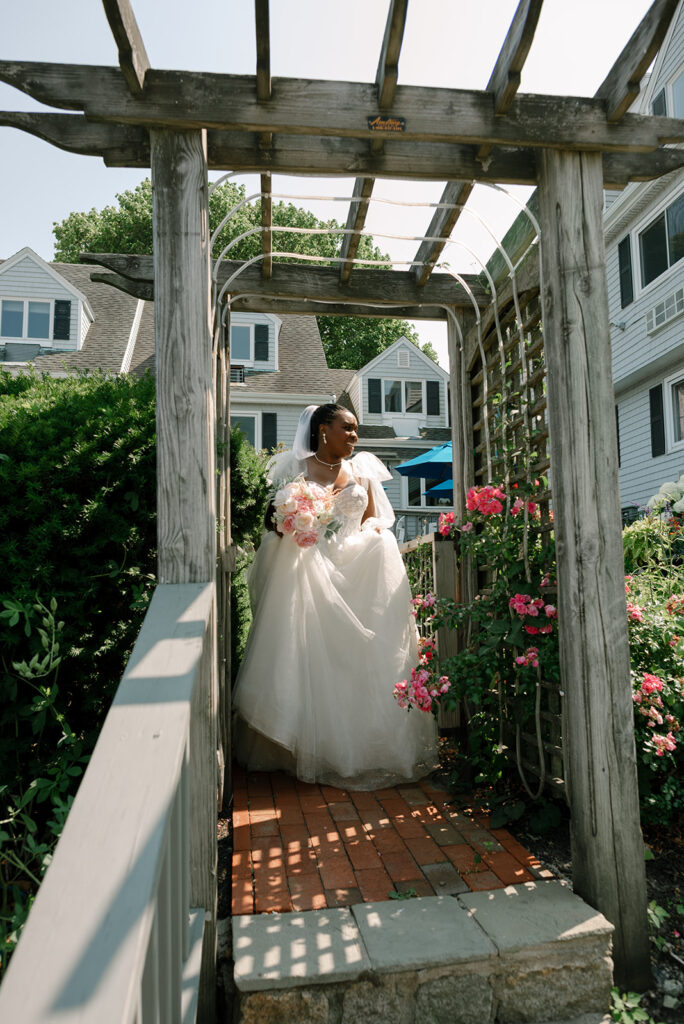 A bride in her wedding gown carries a blush bouquet as she walks under a pergola in the garden at her New England wedding at Barker House in Scituate, MA
