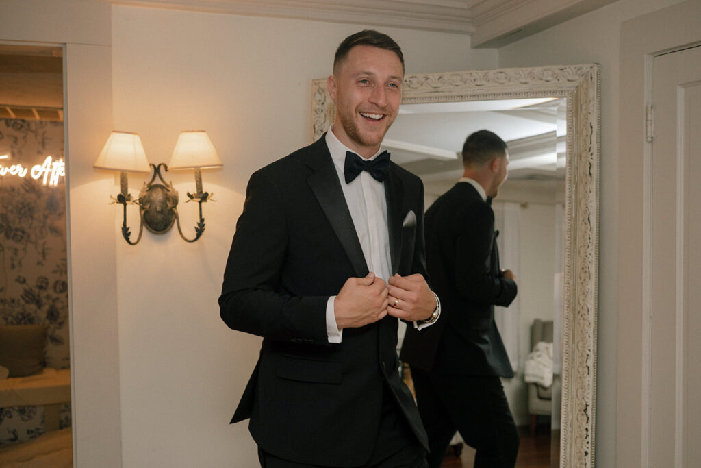 A groom smiles as he adjusts his tux in the getting ready suite at Barker House before his New England wedding ceremony 
