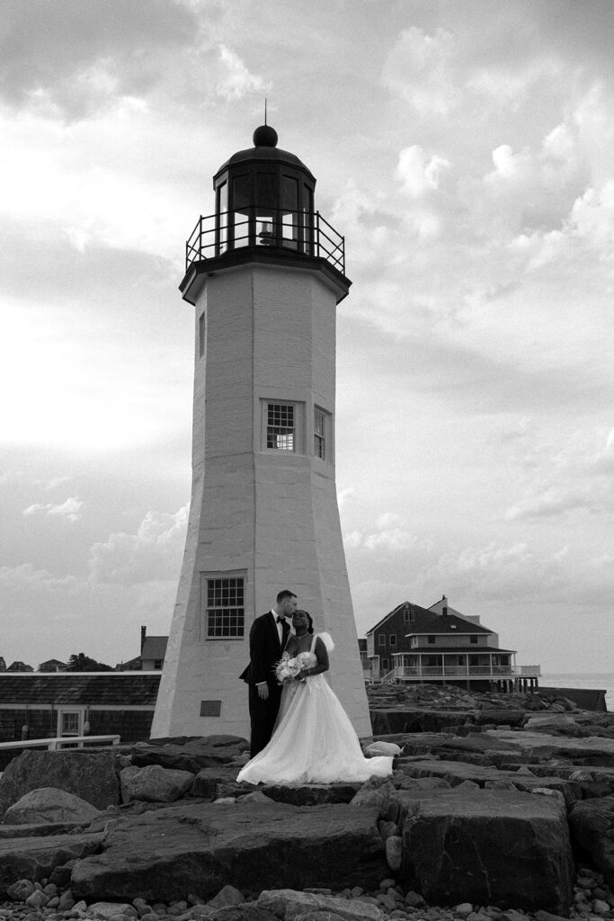 A black and white image showcases newlyweds standing beneath the lighthouse near Barker House by Wedgewood Weddings in Scituate, MA
