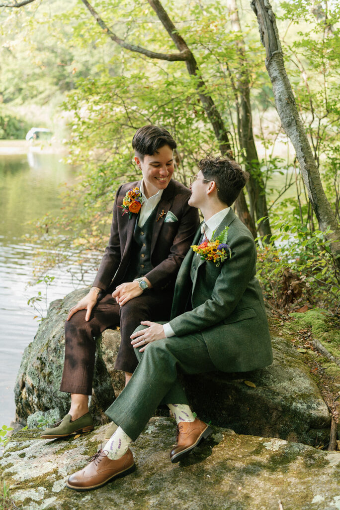 Newlyweds smile seated on the rocky short overlooking the water at their Westchester County wedding